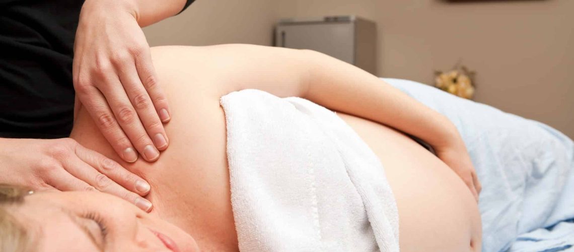 A pregnant woman is receiving a prenatal massage from a female massage therapist. This type of massage is used to relieve tension, stress and discomfort during pregnancy. The background includes a stone/towel warmer on the left, flowers, a blurred accreditation document and a shelf with candles and oils.